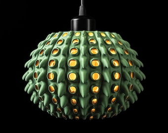 May include: A spherical pendant light fixture with a matte sage green exterior featuring a textured, spiky surface with numerous circular cutouts. Warm yellow light glows from within the openings, and a black cord extends from the top.