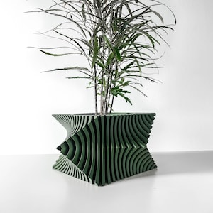 May include: A dark green planter with a complex, layered, and ribbed geometric structure that creates a twisting, hourglass-like silhouette. A tall, thin-leaved plant with slender, dark green stems and pointed foliage grows from the center.
