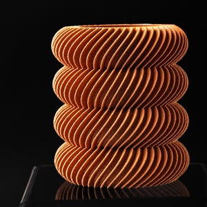 May include: A cylindrical object with a textured, ribbed surface in a warm terracotta orange color. The structure consists of four stacked, rounded segments, each featuring a repeating diagonal pattern that creates a distinct, ridged appearance.