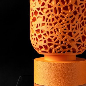 May include: A bright orange cylindrical planter featuring a complex, open-work geometric lattice pattern across its upper section. The base is a solid, textured orange cylinder that supports the intricate, hollowed-out upper body of the vessel.