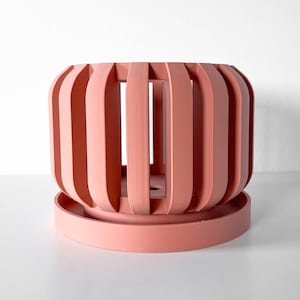 May include: A matte coral-colored planter with a slatted, cage-like design featuring vertical bars and a matching circular drainage saucer base. The structure is open between the slats, revealing a central opening at the bottom of the pot.