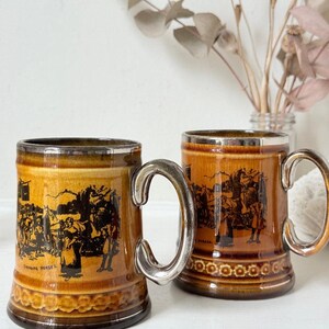May include: Two brown ceramic mugs with a glossy finish and silver-toned rims and handles. Each mug features a black printed scene of people and horses with the text Changing Horses on the front. The base of each mug has a raised circular pattern.