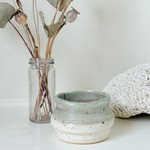 May include: A small, handmade ceramic pot with a two-tone glaze. The top half is a muted seafoam green, transitioning to a speckled white base. The pot has a rounded shape with a textured, ribbed design. A clear glass jar with dried stems and seed pods is in the background.
