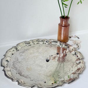 May include: A round silver-toned serving tray with a scalloped edge and an intricate embossed floral pattern across its surface. The tray features a reflective metallic finish and a slightly tarnished appearance, showcasing detailed scrollwork.