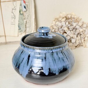 May include: A round ceramic lidded jar featuring a glossy black base glaze with a decorative drip pattern of light blue glaze cascading from the rim and the lid handle. The vessel has a wide, bulbous body and a matching circular lid with a knob.