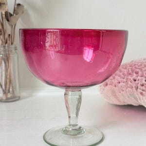 May include: A vibrant magenta glass goblet featuring a wide, rounded bowl and a clear, slender stem that connects to a circular base. The glass has a smooth, polished finish with subtle variations in colour intensity throughout the bowl structure.
