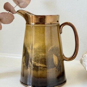 May include: A ceramic jug with a metallic bronze rim and handle, featuring a gradient glaze that transitions from light olive green to dark brown. The body displays a printed landscape scene with a sailboat and the text Cromarty.