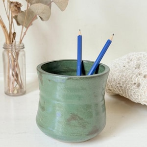 May include: A cylindrical ceramic cup with a matte sage green glaze and subtle brown undertones. The cup has a slightly flared rim and a rounded base. Two blue pencils are placed inside the cup, extending upwards from the opening.