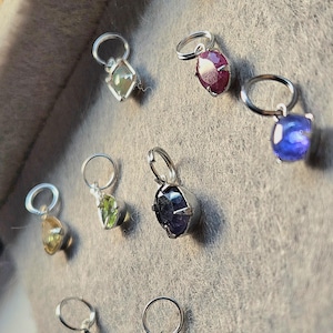 May include: A collection of gemstone pendants in various colors, including purple, red, green, yellow, and blue. Each pendant features a unique, faceted gemstone set in a silver-colored metal setting with a small loop for a chain. The pendants are displayed against a gray background.