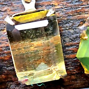May include: A rectangular pendant with a clear, yellow-toned gemstone. The gemstone is set in a silver-colored metal frame and has a white rectangular accent. The pendant hangs from a silver-colored bail. The background is a dark, textured surface.