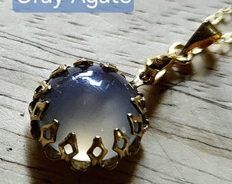 Op de afbeelding: Een goudkleurige hangerketting met een grijze agaatsteen. De steen is gezet in een decoratieve gouden setting met een kroonontwerp. De tekst "Gray Agate" staat in een blauwe rechthoek boven de hanger.