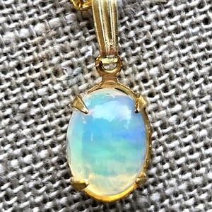 May include: A gold pendant necklace featuring an oval opal gemstone. The opal displays a play of color with hues of blue, green, and yellow. The pendant is suspended from a delicate gold chain, showcasing a classic jewelry design.
