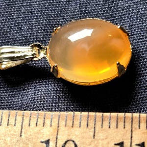 May include: A gold pendant featuring an oval, amber-hued gemstone. The gemstone is secured in a gold bezel with four prongs. The pendant includes a gold bail for a chain. The backdrop is a dark blue fabric.
