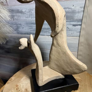 Op de afbeelding: Abstracte sculptuur van twee figuren, één staand en één knielend, gemaakt van beige keramiek. Het beeld staat op een zwarte basis.