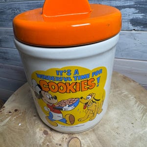 May include: A white ceramic cookie jar with an orange lid. The jar features a cartoon image of Mickey Mouse and Pluto with the text "It's a Wonderful Time for Cookies!"