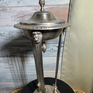 May include: A silver decorative stand with a round top and three legs. The stand features lion head accents and a finial on top.