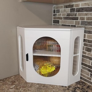 May include: White corner cabinet with a round glass door and vertical side accents. The cabinet is mounted on a countertop and has a black handle. The interior is visible through the glass door.
