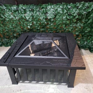 May include: A square, black metal fire pit with a mesh screen and a decorative stone-like pattern around the edges. The fire pit has a rectangular base and is filled with wood. A handle is visible on the side.