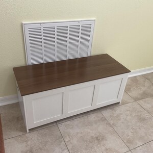 May include: A white wooden storage chest with a dark brown top. The chest has three recessed panels on the front and is positioned against a wall with a white vent.