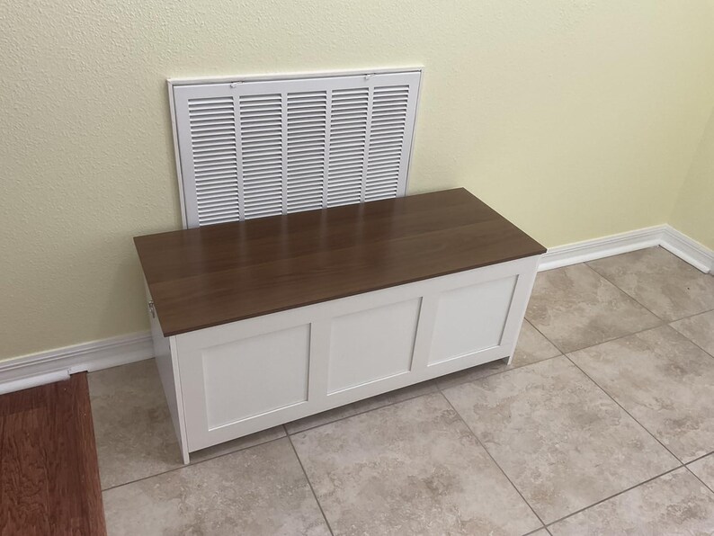 May include: A white wooden storage chest with a dark brown top. The chest has three recessed panels on the front and is positioned against a wall with a white vent.