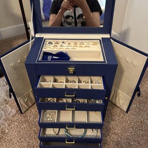May include: A navy blue jewelry box with a mirrored lid and multiple drawers. The box is open, revealing various rings, bracelets, and necklaces. The interior is lined with a light beige fabric. Gold-colored hardware accents the drawers.