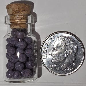 May include: A small clear glass bottle with a cork stopper filled with tiny purple and white swirled spherical beads, positioned next to a United States dime for scale to show the miniature size of the beads contained within the glass vessel.