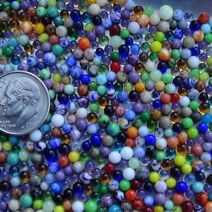 May include: A large collection of colorful glass marbles, some with swirls and patterns, next to a US dime coin. The marbles are various shades of blue, green, red, yellow, and white.