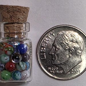 May include: A small glass bottle filled with colorful marbles, capped with a cork. Next to the bottle is a U.S. dime coin, showing the profile of Franklin D. Roosevelt and the year 2021. The marbles are various colors.