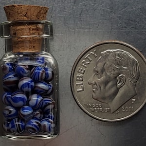 May include: A small clear glass bottle with a cork stopper filled with tiny blue, white, and brown swirled glass beads, positioned next to a United States dime for scale to show the miniature size of the beads and the bottle.