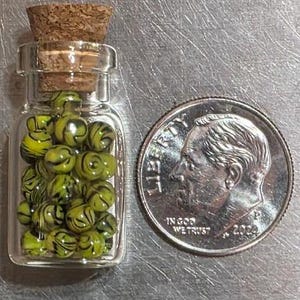May include: A small clear glass bottle with a cork stopper filled with tiny yellow and black swirled beads, positioned next to a United States dime for scale, showing the beads are approximately 0.15 inches in diameter.