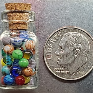 May include: A small glass bottle filled with colorful marbles, corked at the top. A U.S. dime is next to the bottle for size comparison. The marbles are various colors, including blue, green, orange, and yellow.