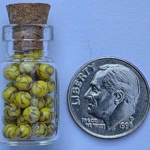 May include: A small glass bottle filled with yellow and brown swirled beads, next to a U.S. dime coin. The bottle has a cork stopper. The dime shows a profile of a man and the words "IN GOD WE TRUST" and the year 1996.
