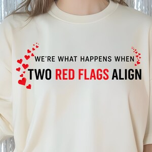 Puede incluir: Camiseta color crema con el texto "WE'RE WHAT HAPPENS WHEN TWO RED FLAGS ALIGN" en rojo y negro, con detalles de corazones rojos. La camiseta tiene mangas cortas y un corte holgado.