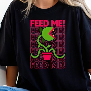 Puede incluir: Camiseta negra con un diseño de planta de dibujos animados y el texto rosa "FEED ME!". La planta tiene una boca grande y abierta y está en una maceta rosa. El texto se repite verticalmente. La camiseta es de corte holgado.