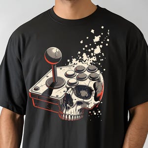 May include: Black t-shirt featuring a graphic of a skull with a joystick design. The joystick has a red outline and a gray and black color scheme. The skull has a white and gray color scheme. The shirt is a crew neck.