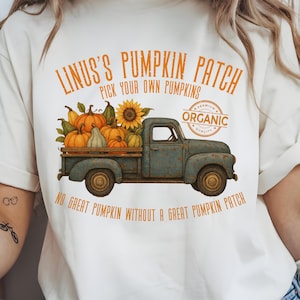 Può includere: T-shirt color crema con un design di un camioncino blu vintage pieno di zucche e girasoli. Il testo "LINUS'S PUMPKIN PATCH" e "PICK YOUR OWN PUMPKINS" sono stampati sopra il camion. La parola "ORGANIC" è anch'essa stampata.