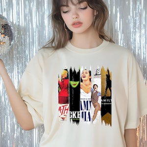 May include: Cream-colored t-shirt featuring a collage of musical theater posters. The design includes images and text from various Broadway shows, such as "Wicked" and "Hamilton". The shirt has a crew neck and short sleeves.