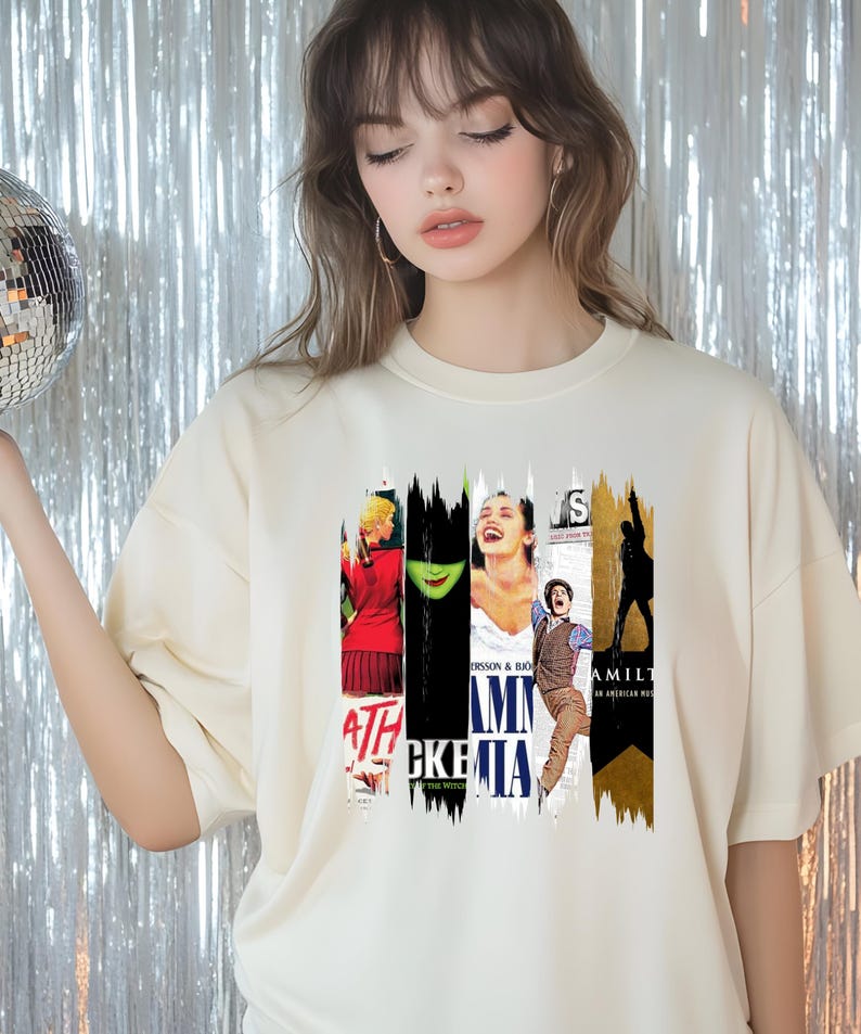 May include: Cream-colored t-shirt featuring a collage of musical theater posters. The design includes images and text from various Broadway shows, such as "Wicked" and "Hamilton". The shirt has a crew neck and short sleeves.