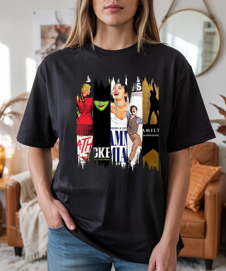 May include: Black t-shirt featuring a collage of musical theater posters. The design includes images and text from popular Broadway shows such as "Wicked," "Mamma Mia," and "Hamilton." The shirt is a crew neck and has short sleeves.