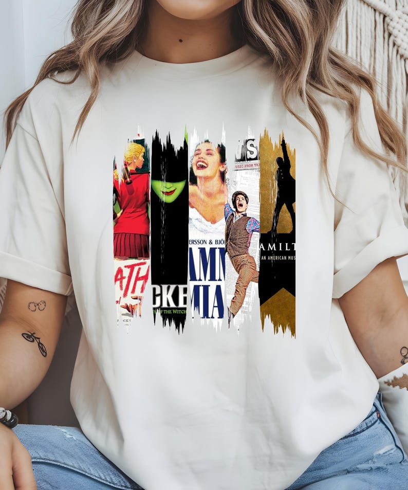 May include: Cream-colored t-shirt featuring a collage of musical theater posters. The design includes images and text from various shows, including "Wicked," "Mamma Mia," and "Hamilton." The shirt has a classic crew neck and short sleeves.