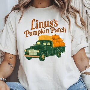 Può includere: T-shirt color crema con un camioncino verde vintage carico di zucche e fieno. Il testo "Linus's Pumpkin Patch" è arcuato sopra il camion in lettere marroni e arancioni. Una maglietta grafica a tema autunnale.