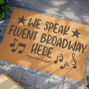 May include: A rectangular doormat with the text "WE SPEAK FLUENT BROADWAY HERE" in dark brown. Musical notes and stars accent the text. The phrase "Enter With Caution" is printed below the main text. The mat is tan and sits on a stone surface.