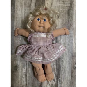 Op de afbeelding: Een zachte pop met blond krullend haar, blauwe ogen en kuiltjes in de wangen. De pop draagt een lichtpaarse en witte gingham jurk met korte ruche mouwen en twee horizontale witte lintstrepen rond de zoom van de rok.