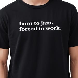 May include: Black t-shirt with the white text "born to jam. forced to work." The shirt is a crew neck style and appears to be made of a soft material. The text is centered on the chest.