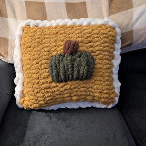 May include: A square, hand-knitted throw pillow featuring a mustard yellow base with a white border. Centered on the pillow is a dark green, textured pumpkin shape with a small brown stem, all crafted from thick, chunky yarn in a soft, looped stitch.