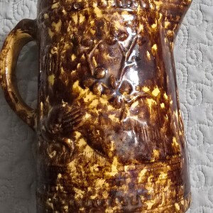 May include: A brown and yellow glazed stoneware pitcher with a floral design and a handle. The pitcher has a mottled glaze and a brown rim.