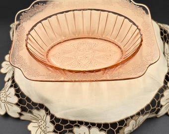 May include: Pink Depression glass serving dish set. The set includes a square plate with floral detailing around the edges and an oval bowl with vertical lines. The bowl sits on top of the plate. The glass has a delicate, translucent appearance.