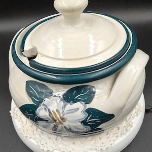 May include: Cream-colored ceramic teapot with a lid and a handle. The teapot is decorated with a floral design in shades of blue, green, and gold. A dark teal band accents the lid. The teapot sits on a white plate with a lace doily.