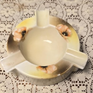 May include: A round ceramic ashtray featuring a cream-colored center and a wide rim decorated with soft yellow and brown gradients. Three evenly spaced notches are integrated into the rim, which is adorned with delicate painted peach-colored flowers.