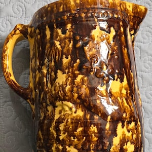 May include: A brown and yellow glazed ceramic pitcher with a mottled design. The pitcher has a wide mouth and a handle.